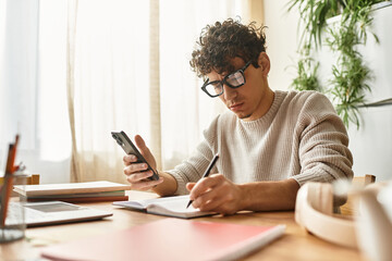Creative young man engaged in productive task at stylish workspace with notebook and phone