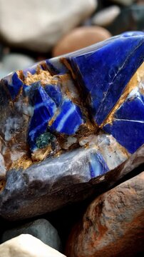 Close-up showcases a cobalt blue mineral formation with white striations and crystalline structure amongst mottled grey, brown, and tan river stones.