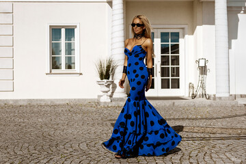 Elegant woman in long polka dot dress poses in front of white palace on sunny summer day. Summer...