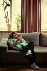 Teenage girl sitting on couch in relaxed pose and using her smartphone. Modern youth lifestyle,...