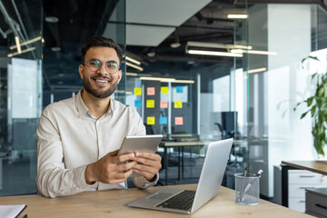 A smiling programmer holding a tablet sits at a desk with a laptop. He looks happy and content in his modern office setting