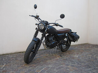 Vista frontale moto custom cafe race nera opaca stile vintage con sella marrone in pelle e fanale anteriore rotondo moto con griglia di protezione vintage