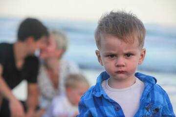 portrait of serious unhappy toddler looking from under his brows by the ocean with his happy family...