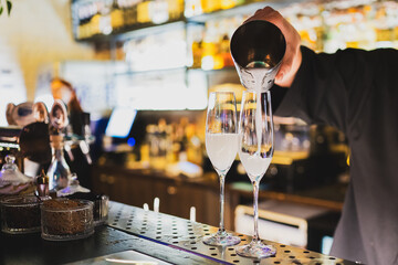 bartender elegantly pours a creamy white cocktail from a shaker into two champagne flutes at a...
