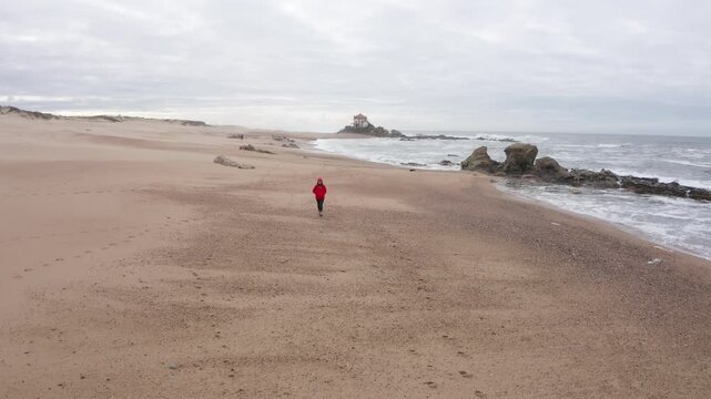Lonely pilgrim woman on Camino de Santiago Portuguese way. Capela do Senhor da Pedra church on sandy beach washed by cold Atlantic ocean waves o, Portugal. Traveling and active people drone 4K video