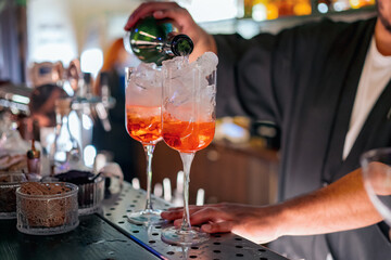 A bartender pours a vibrant orange cocktail into two ice-filled glasses at a bar, capturing the lively atmosphere of nightlife and drink service.