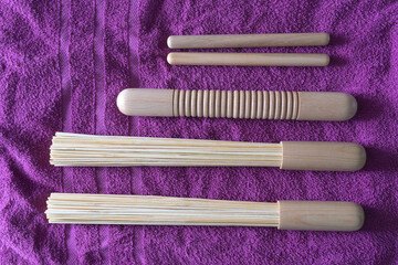 A neatly arranged collection of bamboo massage tools lies on a soft purple towel, including two bamboo whisks, two slender bamboo sticks, and a textured bamboo roller.
