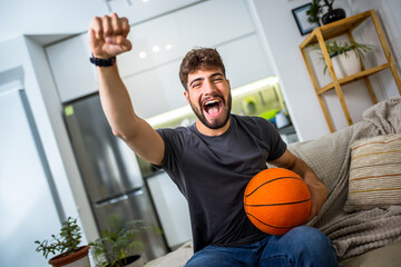 A young man is sitting in his living room, fully engrossed in a basketball game. With a basketball...