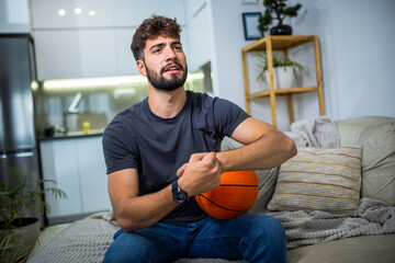 A young man is sitting in his living room, fully engrossed in a basketball game. With a basketball...
