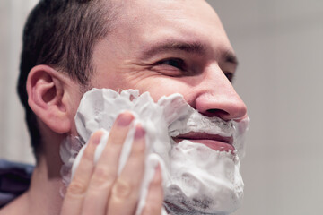 A man is applying shaving foam to his face in front of a bathroom mirror He wears a hooded sweater...
