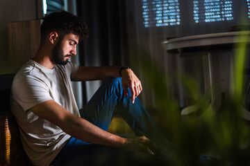 A young man sits alone in a dark room, his face heavy with sadness and his posture reflecting...