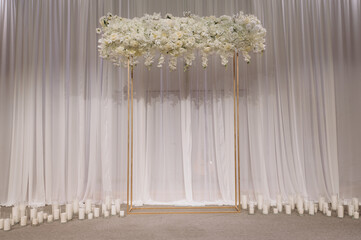 An elegant white arch decorated with fresh flowers, fabrics and decorative elements is located in...