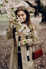 A fragile girl in a beige trench coat with a red handbag, smelling the delicate pink cherry...