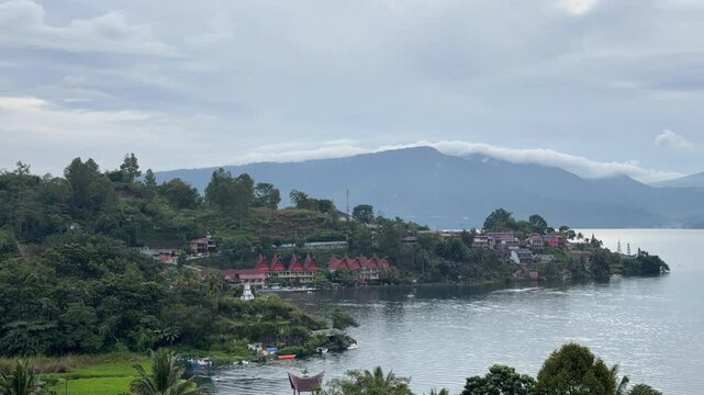 Timelapse of traditional Batak-style buildings and tropical landscape along Lake Toba&rsquo;s shoreline in North Sumatra, Indonesia, under cloudy skies with soft light and calm waters.