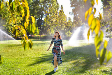 A happy young woman walks through a sunny park where sprinklers are watering the grass. She looks joyful, enjoying the refreshing mist