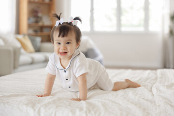 Fototapeta premium Cheerful baby girl crawling on soft bed in bright living room, wearing cute outfit with bows in her hair and smiling at the camera