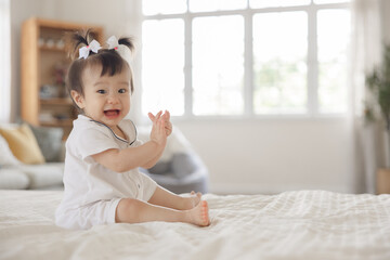 Fototapeta premium Adorable baby girl with playful expression sitting on cozy bed in well-lit room, enjoying joyful moment and clapping hands with happiness