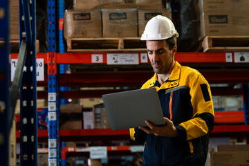 Male warehouse worker wearing yellow uniform and white helmet holding laptop operating inventory...