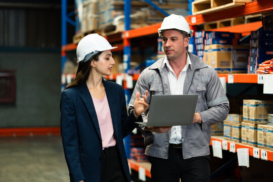 Female logistics manager and male warehouse staff wearing helmets discussing operations with laptop inside warehouse, adult Caucasian team collaborating in inventory control and supply chain workplace