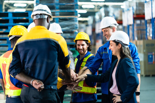 Diverse group of warehouse employees standing in circle placing hands together smiling inside warehouse, male and female workers in helmets and safety uniforms building team spirit in logistics area