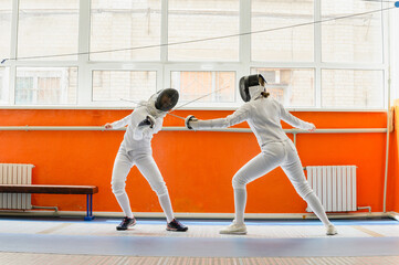 Female fencer striking opponent in bright indoor fencing hall.
