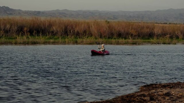 Woman in plaid shirt and beanie paddles a red inflatable packraft on calm spring water at Kapchagay Reservoir, with dry reeds and distant mountains in the background. Captures freedom, adventure, and 