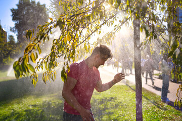 A happy young man stands in a sunny park, smiling with eyes closed as he enjoys the refreshing water spray from active sprinklers