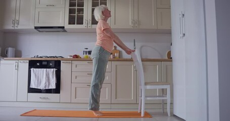Active elderly woman with white hair doing stretching and exercises with chair on orange sports carpet in cozy kitchen at home. Active senior woman