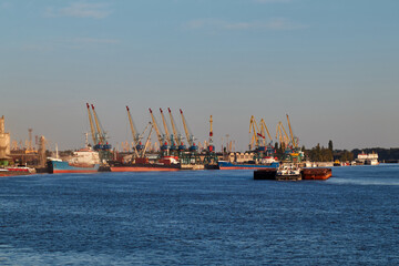 Cargo ships docked at industrial harbor with cranes under clear blue sky. Danube river. Ukraine