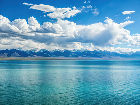 Serene turquoise lake stretches towards snow-capped mountains under a vibrant blue sky adorned with fluffy cumulus clouds.
