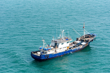 A blue and white fishing boat sails on a calm sea, with gentle waves. The vessel features a deck equipped with fishing gear and a visible wheelhouse.