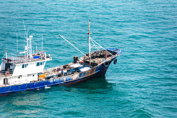 A blue and white fishing boat sails on a calm sea, with gentle waves. The vessel features a deck equipped with fishing gear and a visible wheelhouse. © Mariusz