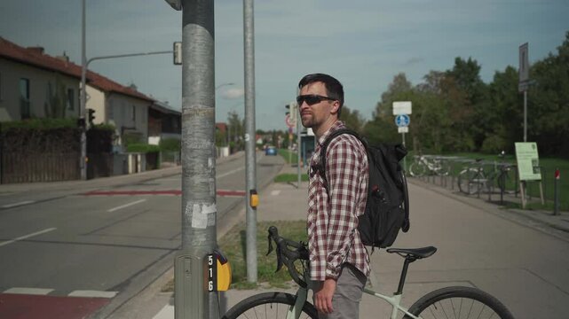 Cyclist wearing plaid shirt and backpack waits at pedestrian crossing in Munich, Germany, pressing a pedestrian push button Anforderungstaster at a traffic light. Urban mobility and bike commuting. 