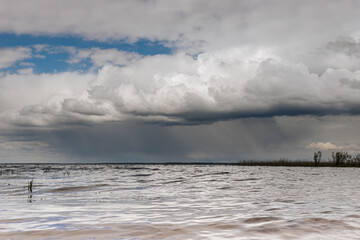 A dark storm approaches over a calm lake, with heavy rain visible on the horizon and dense clouds casting shadows across the sky and water ample copy space.