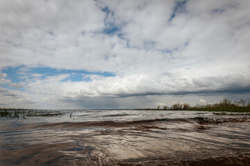 Dramatic clouds loom over a lake shoreline, with distant rainfall visible on the horizon and a hint of blue sky breaking through ample copy space.
