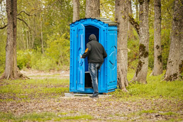 A person wearing a hoodie and jeans enters a blue portable toilet located in a forested area with...