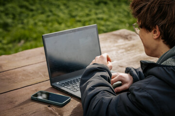 A young man types on a laptop at a wooden outdoor table, wearing a jacket and glasses, with a smartphone beside him and green grass in the background.