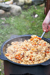 Top view of a Tajik woman cooking hot pilaf in a cauldron over an open fire, stirring the ingredients with a ladle outdoors in a courtyard during summer