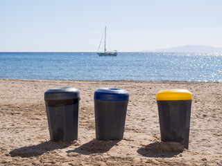 Three trash cans standing on sandy beach with sailboat in background