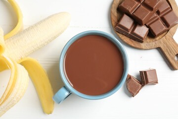 Tasty chocolate milk in cup, pieces and banana on white wooden table, flat lay