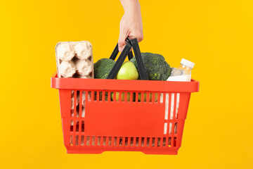 Man holding shopping basket with different products on yellow background, closeup