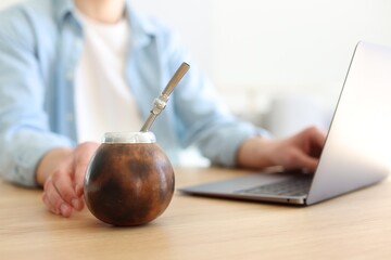Man using laptop at wooden table indoors, focus on traditional yerba mate tea