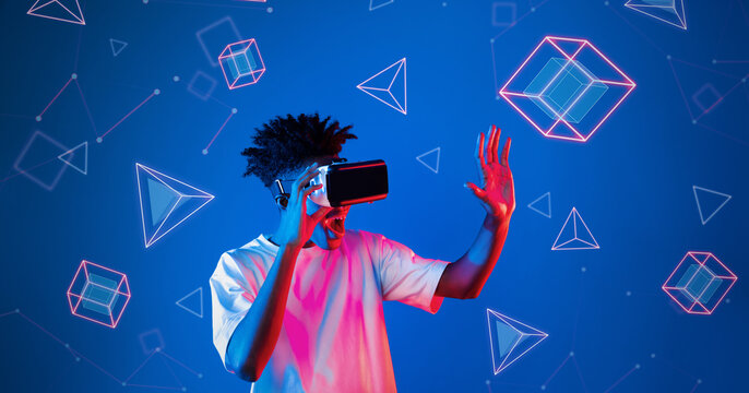 Fototapeta Smart boy standing at blue background while floating geometric shape. African man playing game and wearing VR goggle and casual shirt. Augmented reality technology innovation concept. Ingenuity.