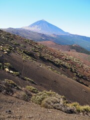 The peak of the Teide volcano against the blue sky and black and red lava formations covered with...
