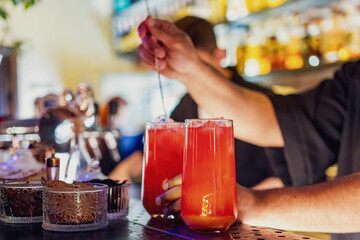 Two tall glasses filled with a vibrant red cocktail are being stirred by a bartender in a lively bar setting, with various bottles and bar equipment in the background.