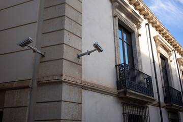 Two security cameras mounted on the corner of an old stone building with wrought iron balconies and...