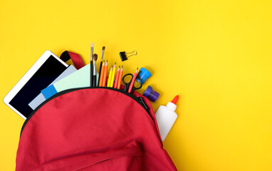 Red school backpack filled with glue, pencils, tablet, brushes, clips and notebooks on yellow. Great image for back to school sales, children&rsquo;s education, and classroom promos.