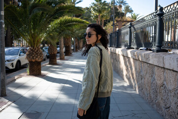 Stylish young woman in sunglasses and casual jacket turning back with a soft smile while walking...