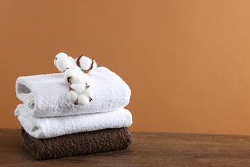 Stack of clean towels and cotton flowers on wooden table against brown background, space for text