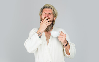 Sleepy yawning man with cup of tea or coffee. Morning refreshment. Sleepy bearded man in bathrobe with cup of hot drink. Tired handsome man with sleepy face trying to wake up with cup of fresh coffee.